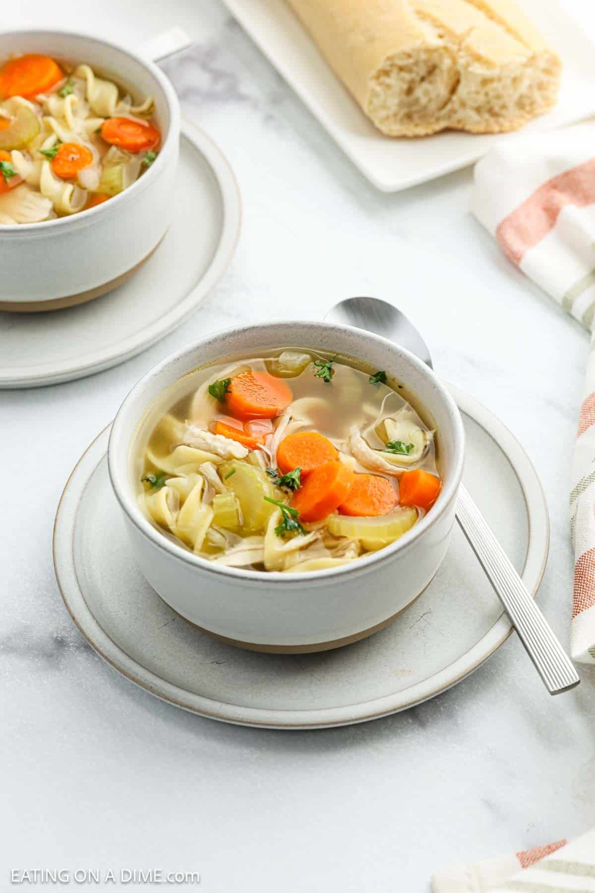 A bowl of Homemade Chicken Noodle Soup with carrots, celery, and parsley sits on a plate with a spoon. In the background, another bowl and a loaf of bread are visible on a white surface.