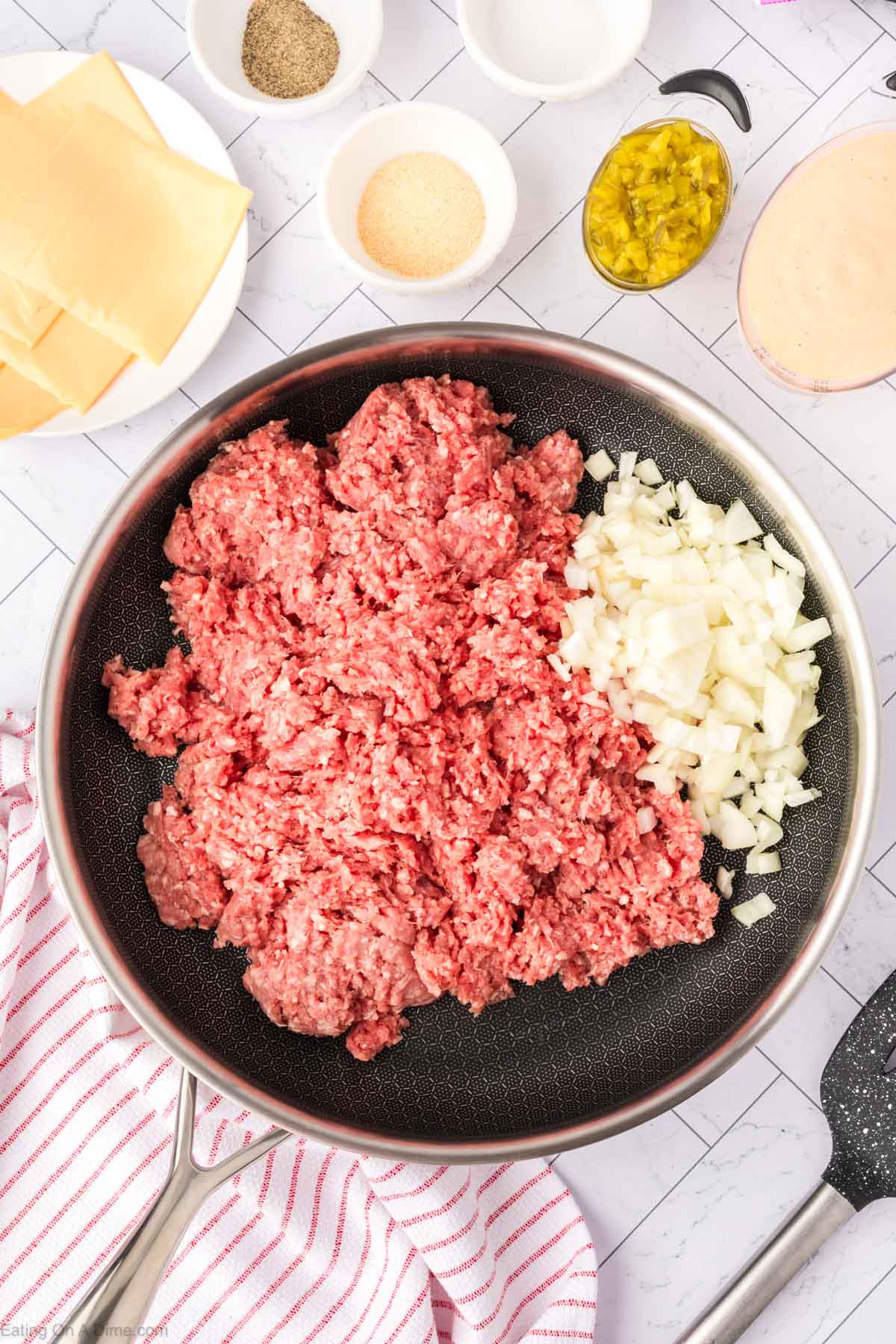 Ground beef in a skillet with diced onions and a plate of slice cheese, small bowls of spices, sauce, and diced pickles