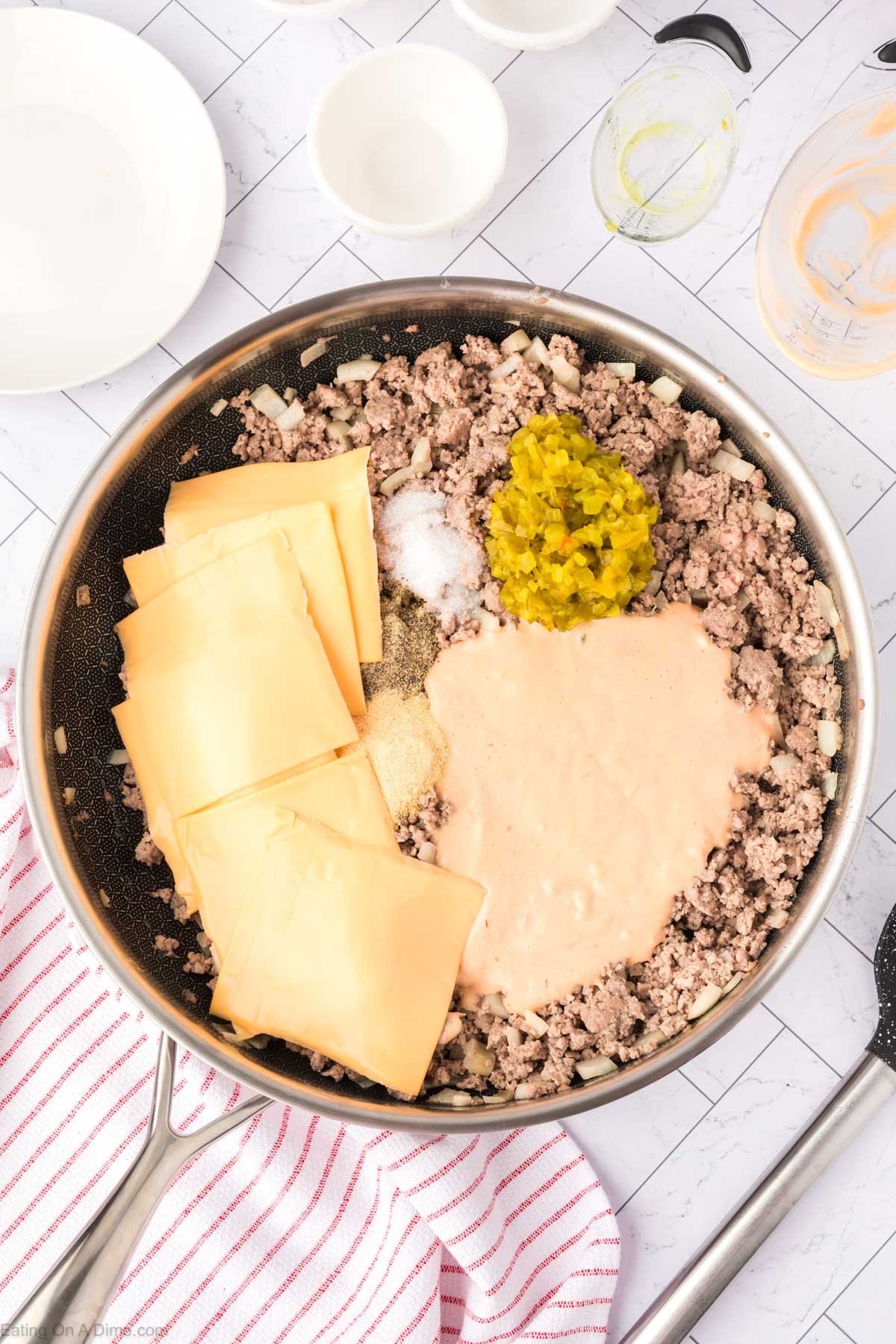 Cooked ground beef and onions in a skillet topped with slice cheese, sauce, diced pickles and spices