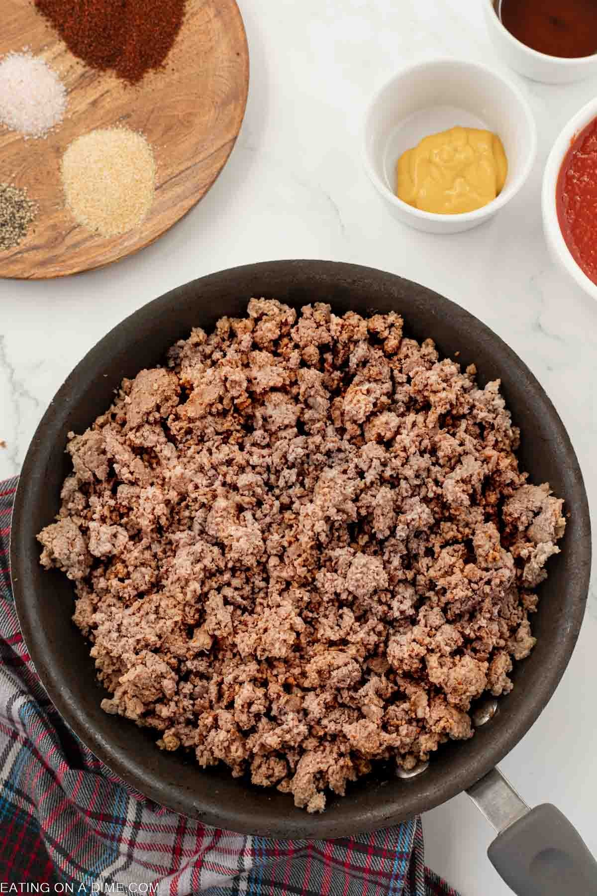 Cooked ground beef in a skillet with a wooden seasoning with small bowls of mustard and ketchup