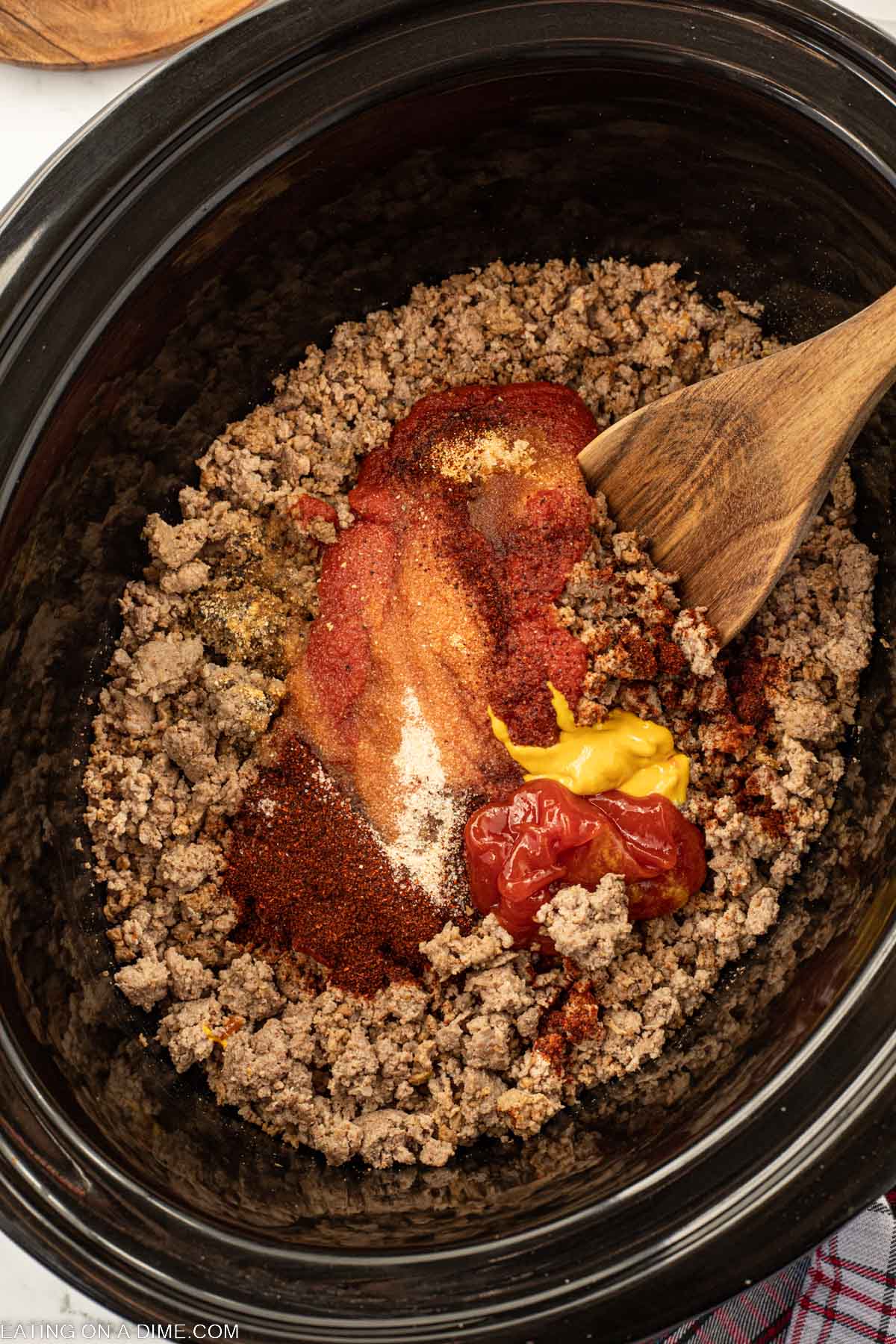 Ground Beef in the slow cooker topped with spices and sauce with a wooden spoon
