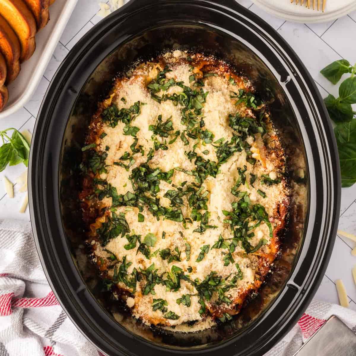 A slow cooker lasagna filled with rich layers, topped with melted cheese and fresh chopped basil, sits on a white countertop beside a towel and a dish of bread rolls.