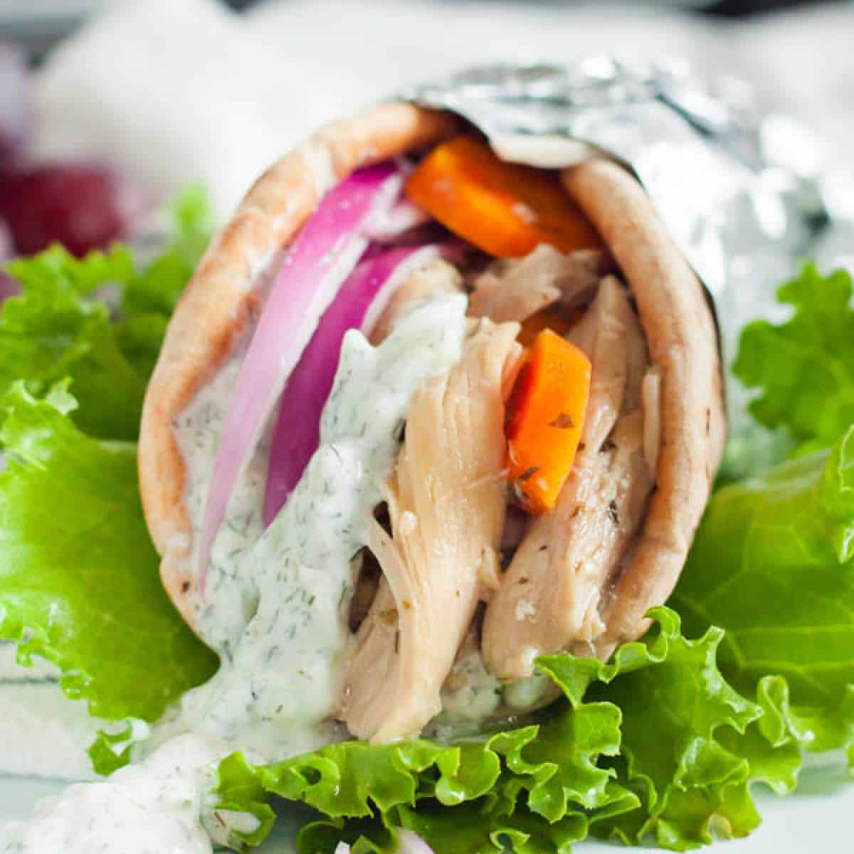 A gyro wrapped in foil with slow cooker chicken gyros, slices of red onion, orange bell pepper, and a drizzle of creamy white sauce. It's presented on a bed of fresh green lettuce.