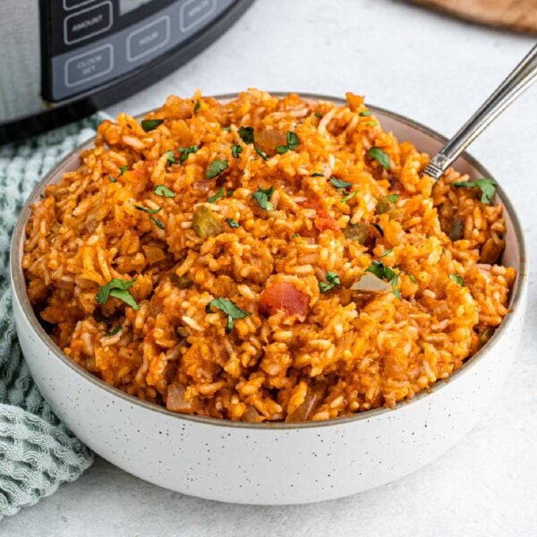 A bowl of crockpot Spanish rice topped with chopped cilantro, featuring visible tomato and green pepper pieces, with a spoon in the bowl and an Instant Pot in the background.