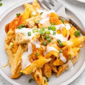 A plate of buffalo chicken pasta bake with pieces of chicken, melted cheese, sliced green onions, and a drizzle of white sauce, served with a fork on the side.