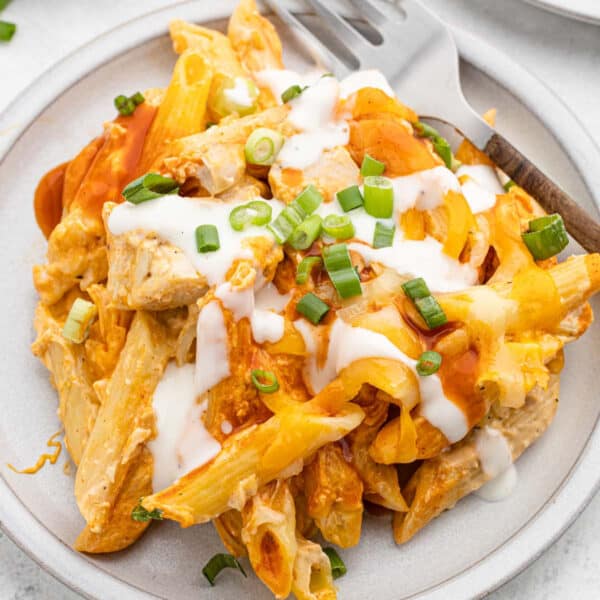 A plate of buffalo chicken pasta bake with pieces of chicken, melted cheese, sliced green onions, and a drizzle of white sauce, served with a fork on the side.