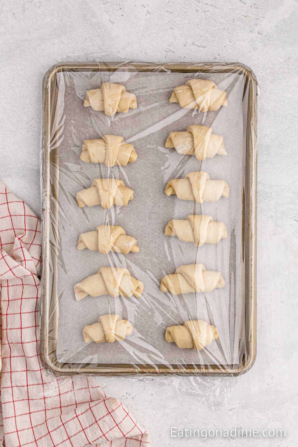 Prepared uncooked crescent rolls on a baking sheet covered with plastic wrap