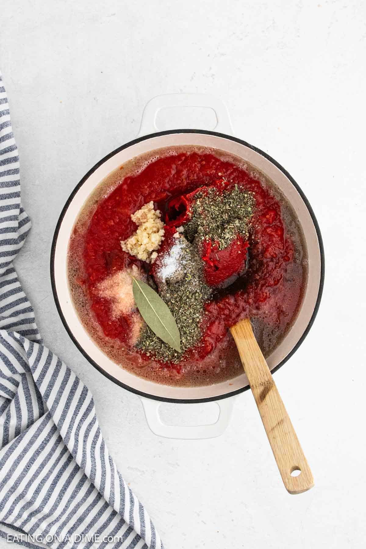 Sauce, minced garlic, spices and a bay leaf in a large pot with a wooden spoon.
