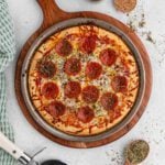 A pepperoni pizza topped with herbs, melted cheese, and a sprinkle of pizza seasoning sits on a round metal tray atop a wooden pizza board, with a pizza cutter and extra seasonings nearby on a light-colored surface.