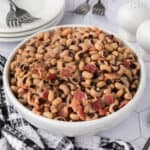 A white bowl filled with a hearty black eyed peas recipe and pieces of bacon, placed on a patterned cloth next to a stack of plates, forks, and salt and pepper shakers.