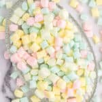A glass platter filled with pastel-colored mini marshmallows and butter mints, mostly in shades of pink, yellow, green, and blue, rests on a marble surface with some sweets scattered around it.