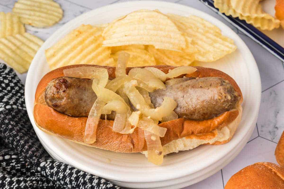 Brats in a bun topped with grilled onions on a plate with potato chips.