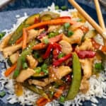A plate of white rice topped with teriyaki chicken stir fry, snap peas, carrots, red bell pepper, zucchini, green onions, and sesame seeds, served with chopsticks.