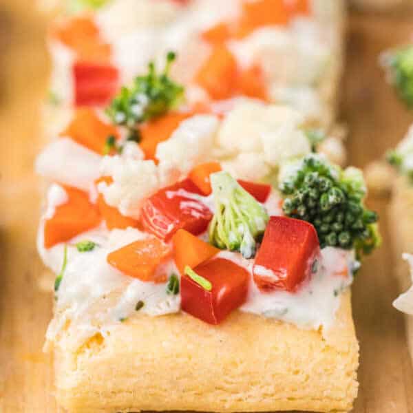 A close-up of a rectangular slice of crescent roll veggie pizza, topped with creamy white spread, diced red bell peppers, broccoli florets, and cauliflower pieces on a wooden surface.
