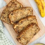 Four slices of banana bread with applesauce are arranged on a wooden cutting board. A yellow banana and a green patterned cloth are nearby, all set on a marble surface.