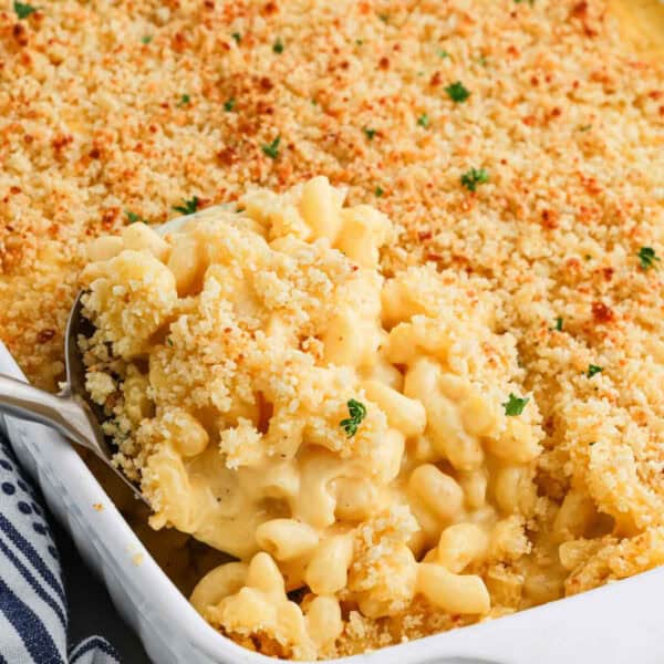 A serving spoon scoops creamy Baked Mac and Cheese with a golden, crispy breadcrumb topping from a white baking dish, garnished with small green herbs.