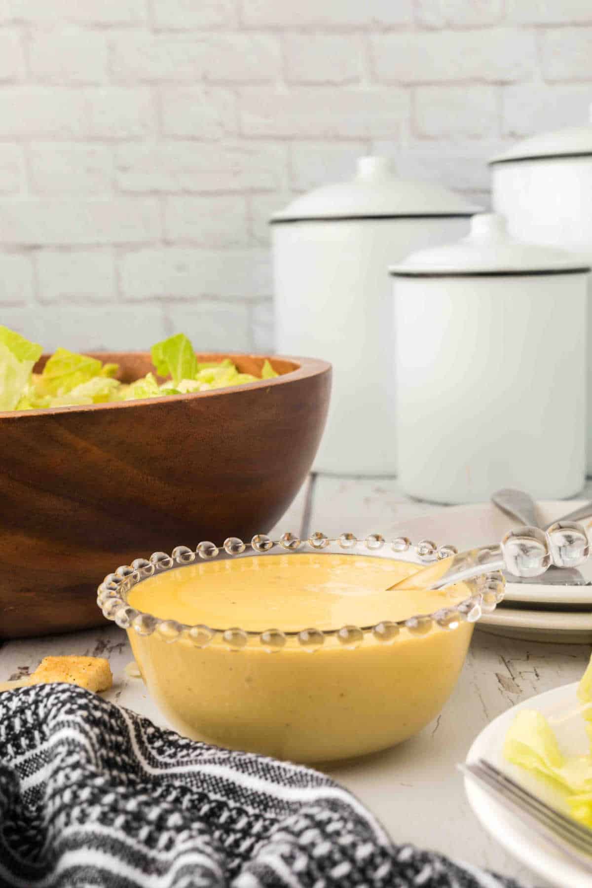 A decorative glass bowl filled with creamy Caesar Salad Dressing, served alongside a large wooden bowl of fresh romaine lettuce with white kitchen canisters in the background.