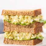 A close-up of a stacked egg salad sandwich, made from with whole wheat bread and leafy lettuce, placed on a white plate.