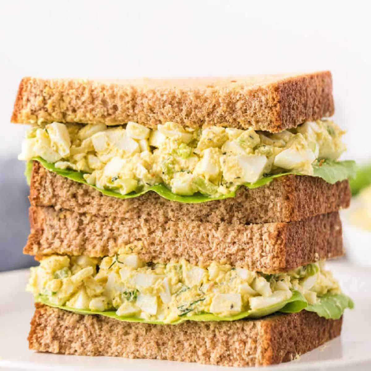 A close-up of a stacked egg salad sandwich, made from with whole wheat bread and leafy lettuce, placed on a white plate.