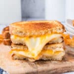 A stack of two grilled cheese sandwiches, inspired by the Starbucks Grilled Cheese Recipe, with golden, crispy bread and melted cheese oozing out, placed on a wooden board with chips in the background.