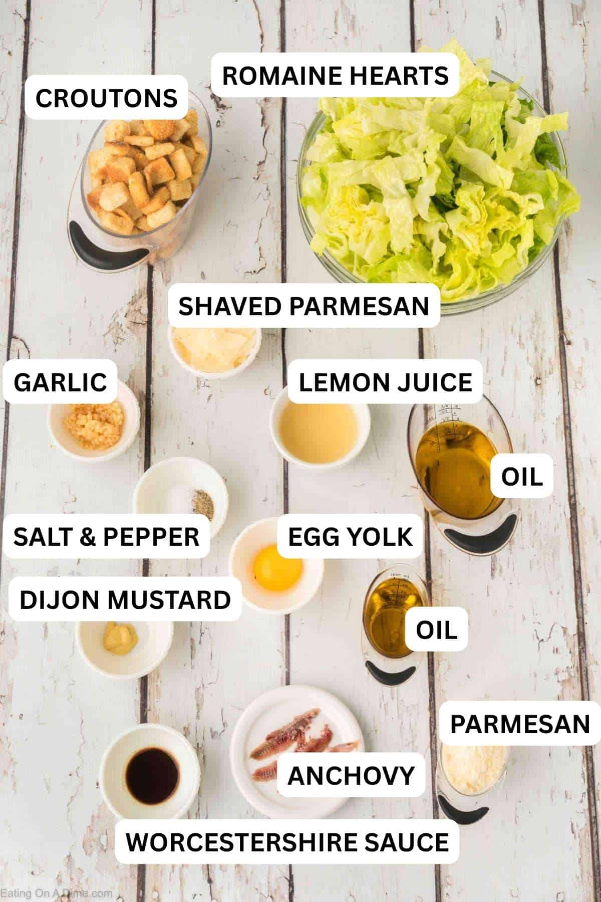 Ingredients needed to make this recipe which includes crisp romaine lettuce, crunchy croutons, shaved parmesan, garlic, anchovies, and an egg yolk, all arranged on a white wood surface.