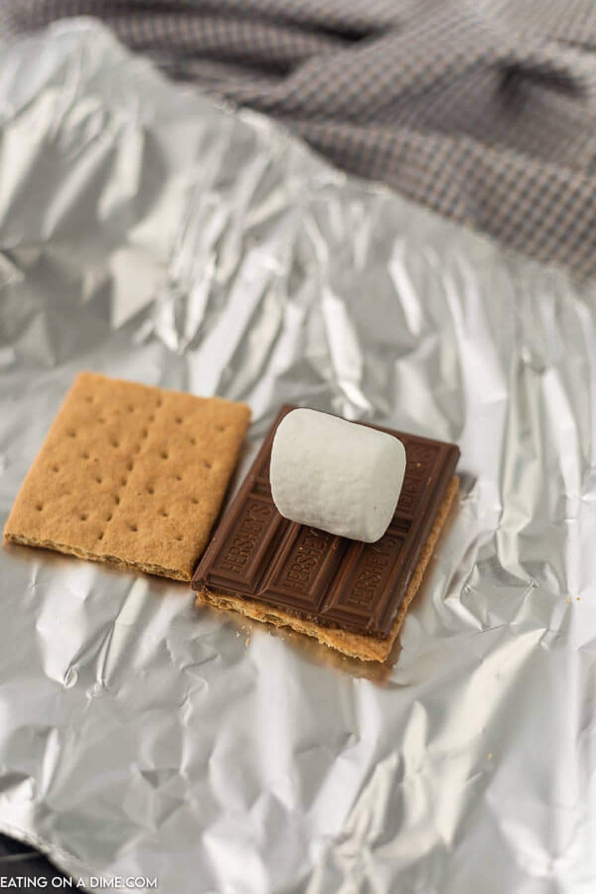 A piece of graham cracker, chocolate and a marshmallow stacked on top of each other on a piece of foil.  