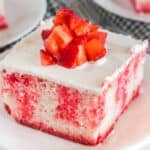 A slice of strawberry Jello Poke Cake topped with diced fresh strawberries and covered in white whipped cream frosting, served on a white plate.