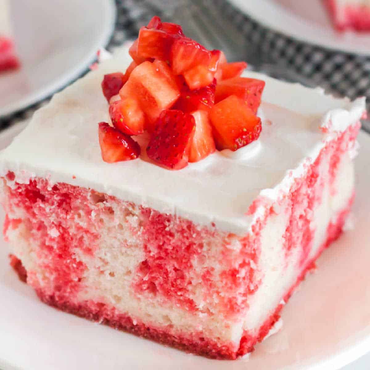 A slice of strawberry Jello Poke Cake topped with diced fresh strawberries and covered in white whipped cream frosting, served on a white plate.