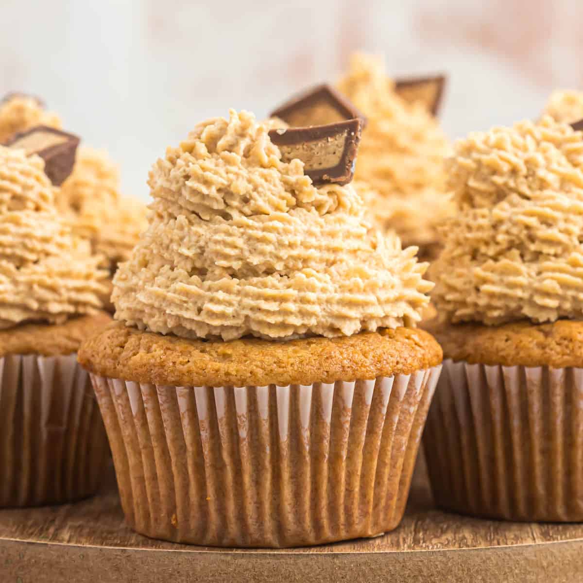 Close-up of peanut butter cupcakes with thick swirls of light brown frosting, topped with chocolate pieces, arranged in a row on a wooden surface against a blurred background.