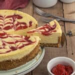 A slice of Strawberry Swirl Cheesecake with a graham cracker crust is being lifted from the whole cake. A bowl of strawberry topping sits nearby on a wooden table, ready to add extra sweetness.