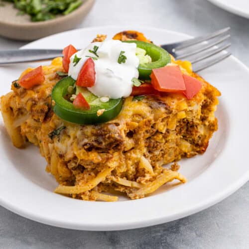 A slice of cheesy Chorizo Breakfast Casserole topped with sour cream, sliced jalapeños, diced tomatoes, and fresh herbs, served on a white plate with a fork.