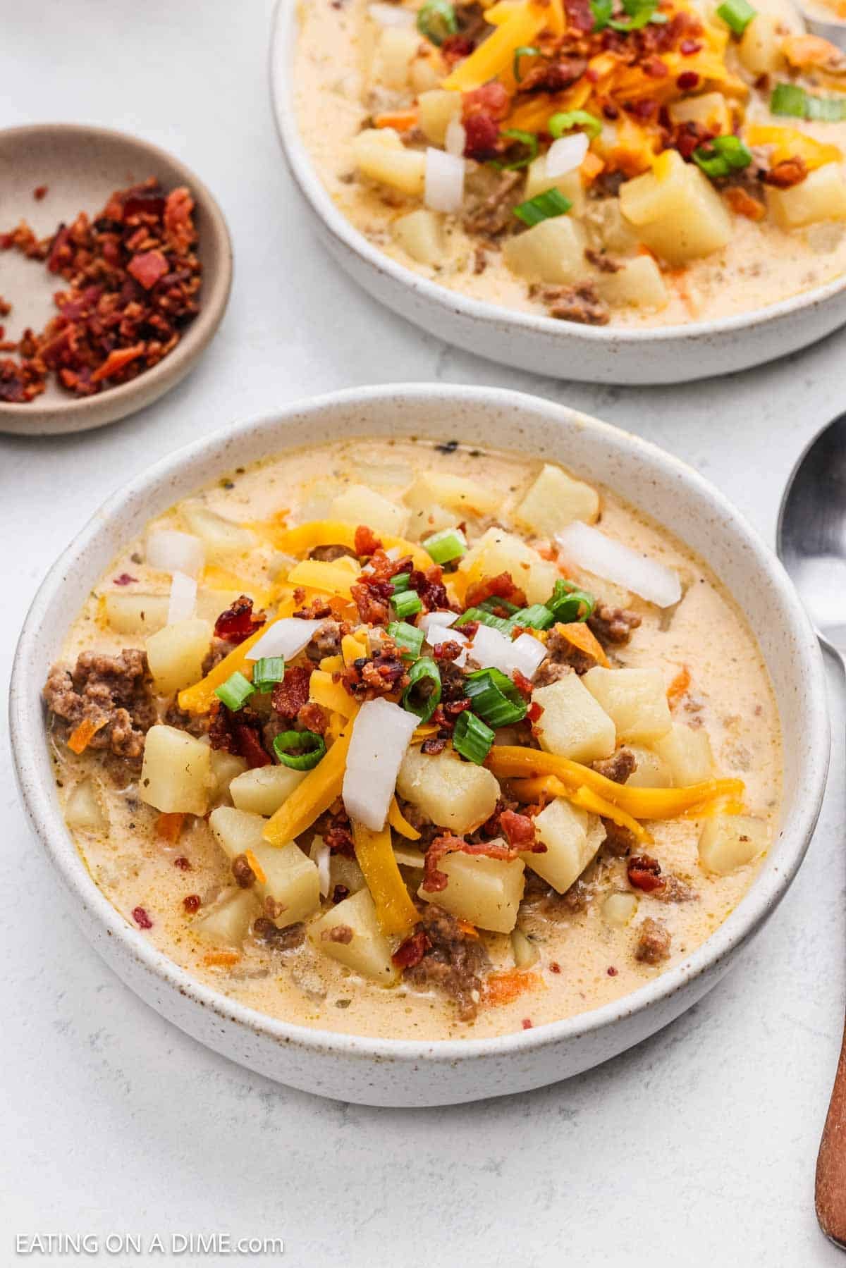 A serving of the potato and ground beef soup in a bowl topped with green onions, shredded cheese and bacon.