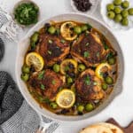 A skillet with four seasoned Moroccan Chicken thighs, garnished with lemon slices, green olives, raisins, and fresh herbs. Small bowls of parsley, raisins, and olives, plus a piece of flatbread sit nearby.