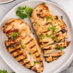 Two grilled chicken breasts on a white plate, one whole and one sliced, showcase visible grill marks and are garnished with chopped parsley. Marinated in Buttermilk Chicken Marinade, they're served with a sprig of fresh parsley for a flavorful finish.