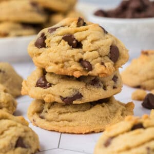 Three Chocolate Chip Walnut Cookies are stacked on top of each other, with more cookies and chocolate chips scattered in the background on a white surface.