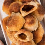A plate piled with golden-brown Resurrection Rolls dusted with cinnamon; some are bitten open, revealing a sweet, brown filling inside.