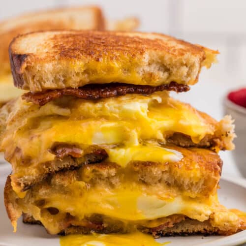 A close-up of a Fried Egg Sandwich cut in half, filled with melted cheese, crispy bacon, and a runny fried egg, with the yolk dripping onto the toasted bread.