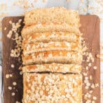 A loaf of Oatmeal Bread, partially sliced, sits on a wooden cutting board with scattered oats around it. The bread has a golden crust and a soft, airy interior.