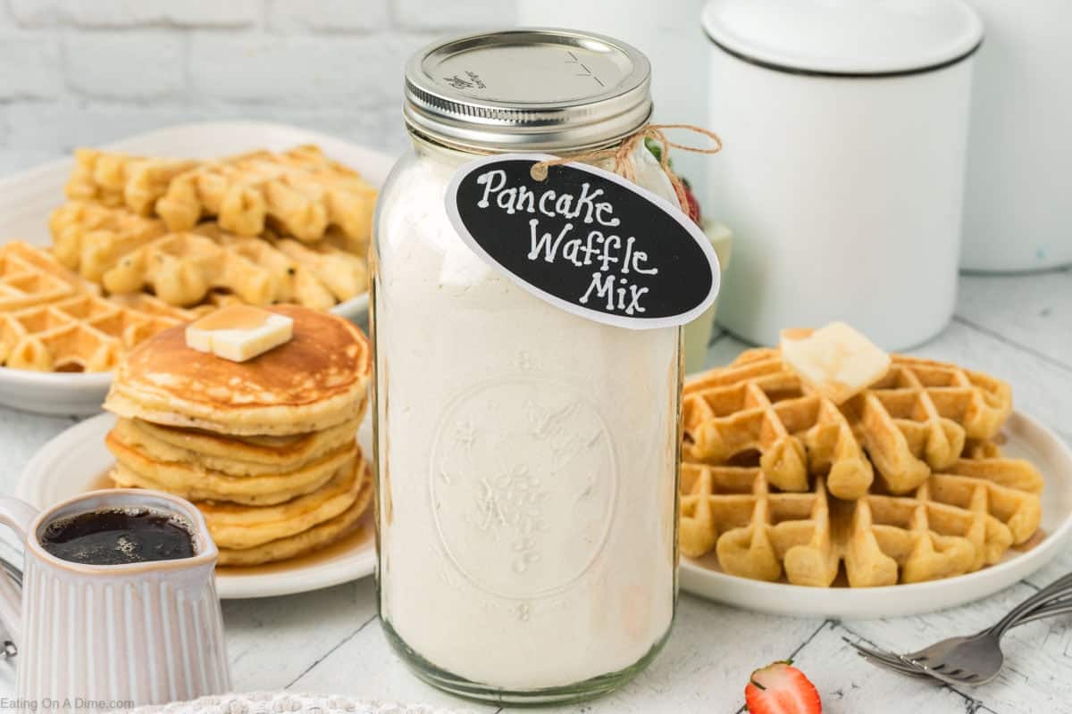 A jar of Pancake Waffle Mix with a black label sits on a table, surrounded by plates of fluffy waffles and pancakes with melting butter, accompanied by a small syrup pitcher and sliced strawberries. A white kitchen canister is visible in the background.