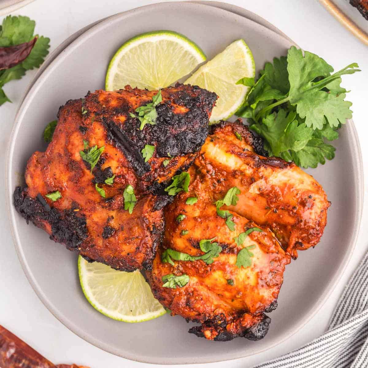 A plate with two pieces of grilled, marinated pollo asado garnished with chopped cilantro, served with lime slices and a sprig of fresh cilantro on the side.