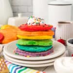 A stack of rainbow pancakes topped with whipped cream and sprinkles sits on a white plate, surrounded by fruit, syrup, and a colorful striped napkin.