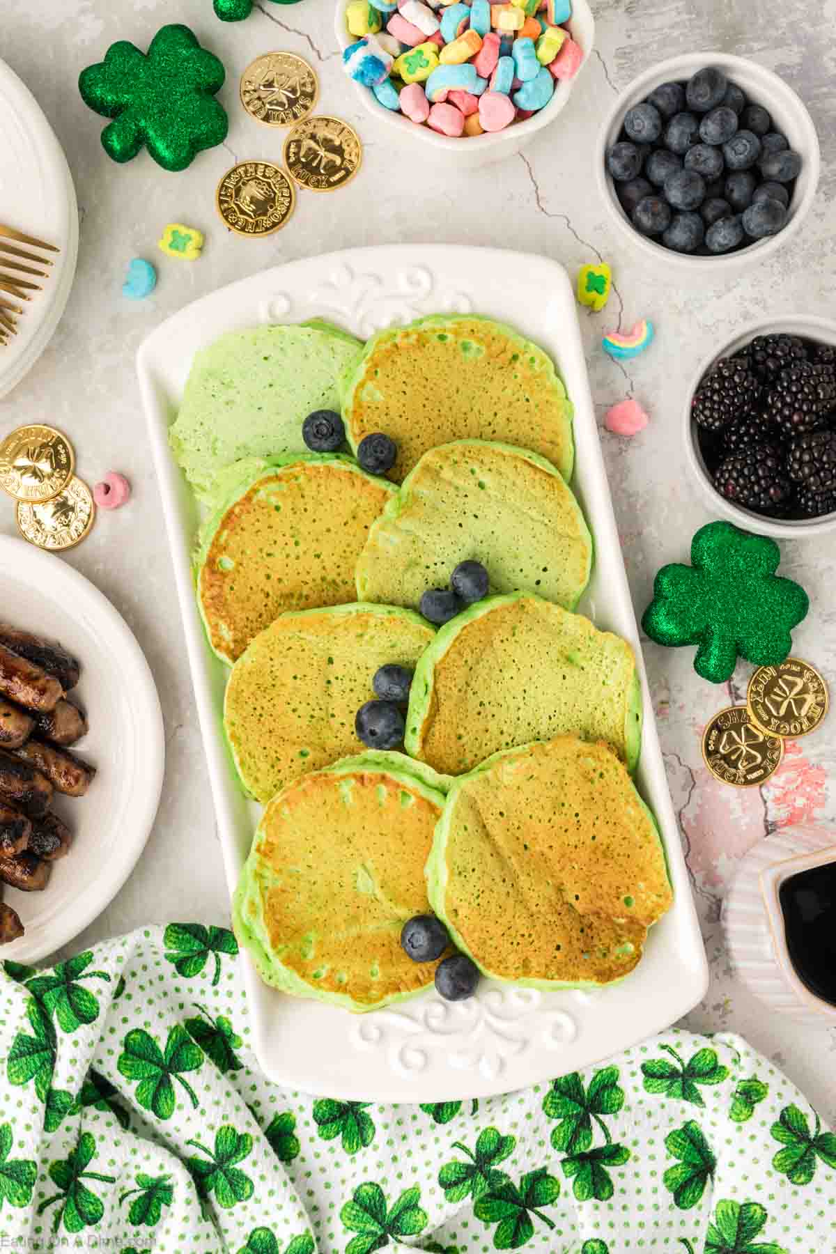Green pancakes stacked on a platter with fresh blueberries and bowls of blueberries and blackberries