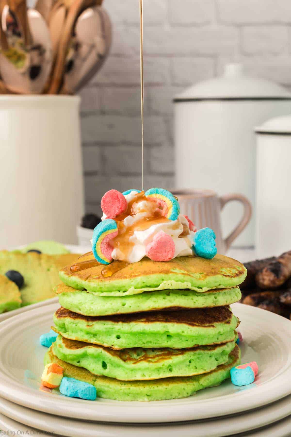 Green pancakes stacked on a plate topped with whipped cream and lucky charms with maple syrup being drizzled on top