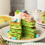 A stack of green St Patrick's Day Pancakes is topped with whipped cream and colorful marshmallow cereal pieces. A wedge is cut from the stack, revealing the fluffy inside. Plates and more pancakes are visible in the background.