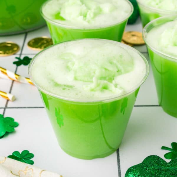 A clear plastic cup filled with bright green, frothy St. Patrick’s Day punch sits on a white surface, surrounded by shamrock decorations, gold coins, and striped straws, creating a festive holiday vibe.