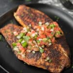 Two pieces of blackened tilapia, inspired by our signature blackened tilapia recipe, are topped with fresh tomato, onion, and cilantro salsa and served on a sleek black plate.