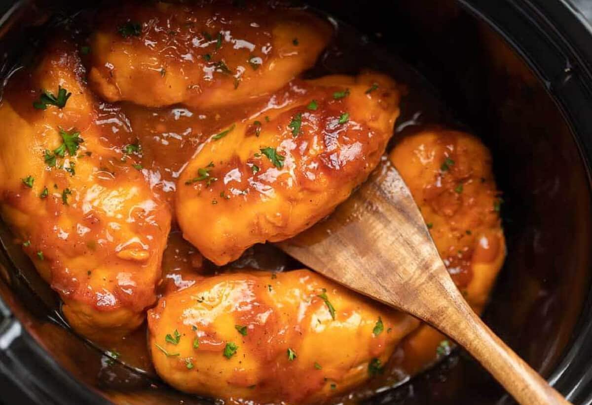 Four pieces of chicken breast covered in a thick, glossy barbecue sauce in a black Crock Pot, garnished with chopped herbs. A wooden spoon rests among the Crock Pot Catalina Chicken pieces.