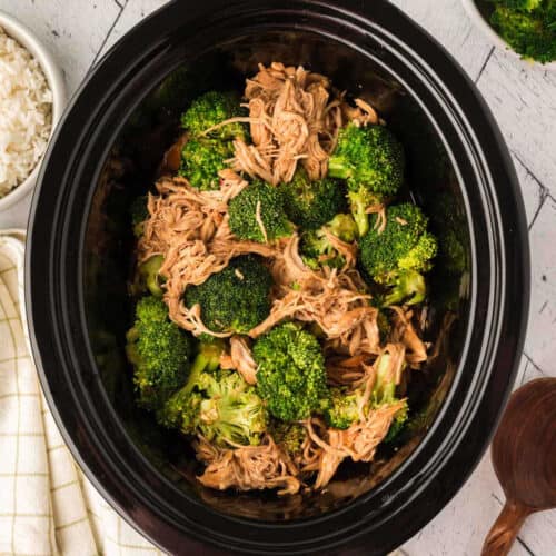 Crock Pot Chicken and Broccoli features shredded chicken and broccoli florets in a black slow cooker, with a bowl of white rice, a wooden spoon, and a plaid napkin nearby on a white surface.