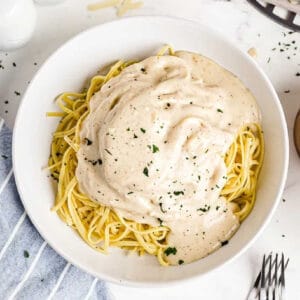 A white bowl of spaghetti topped with Easy Alfredo Sauce and parsley sits on a white surface, surrounded by a fork, salt and pepper shakers, grated cheese, and a striped napkin.