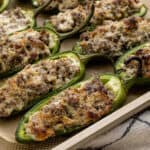 A close-up of Sausage Stuffed Jalapeños shows baked jalapeño peppers halved lengthwise and filled with a browned, cheesy sausage mixture. The filling is bubbly and golden, arranged neatly on a baking tray.